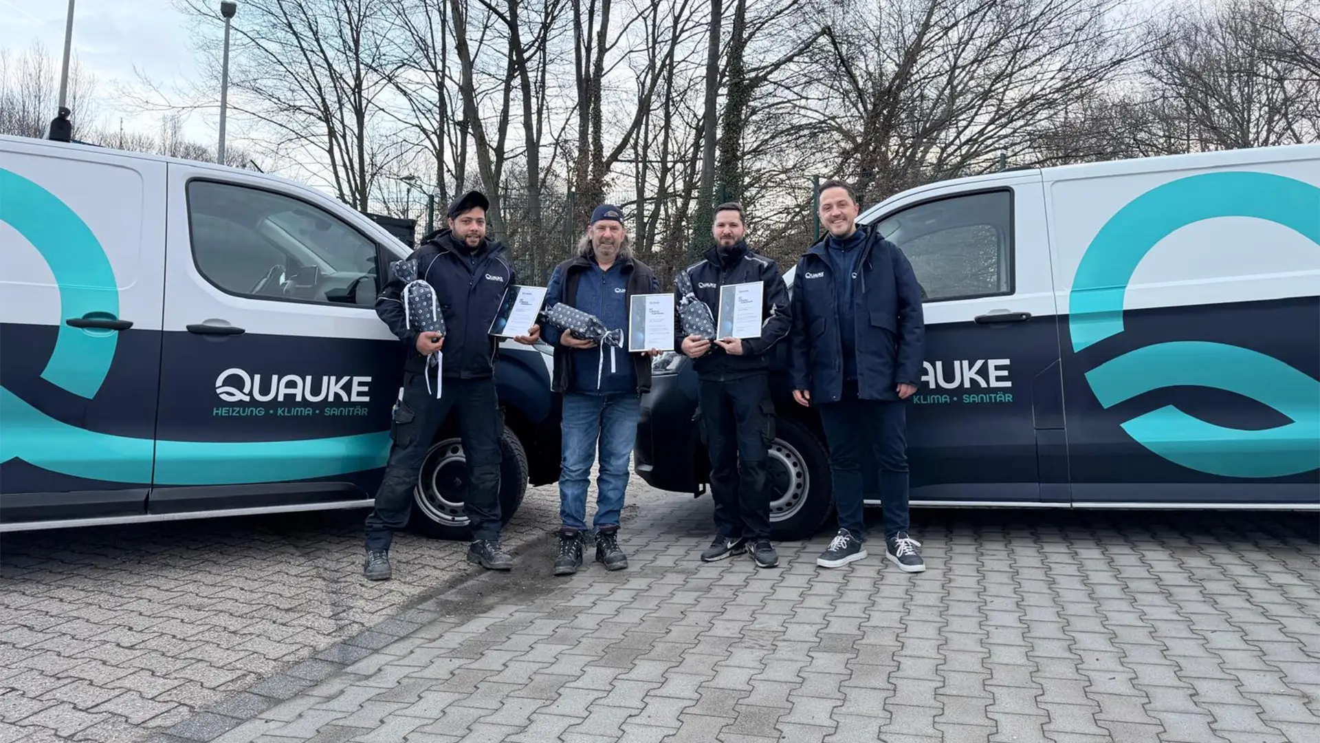 Vier Mitarbeitende stehen zwischen zwei Quauke-Firmenwagen und halten Urkunden sowie kleine Geschenke in den Händen.