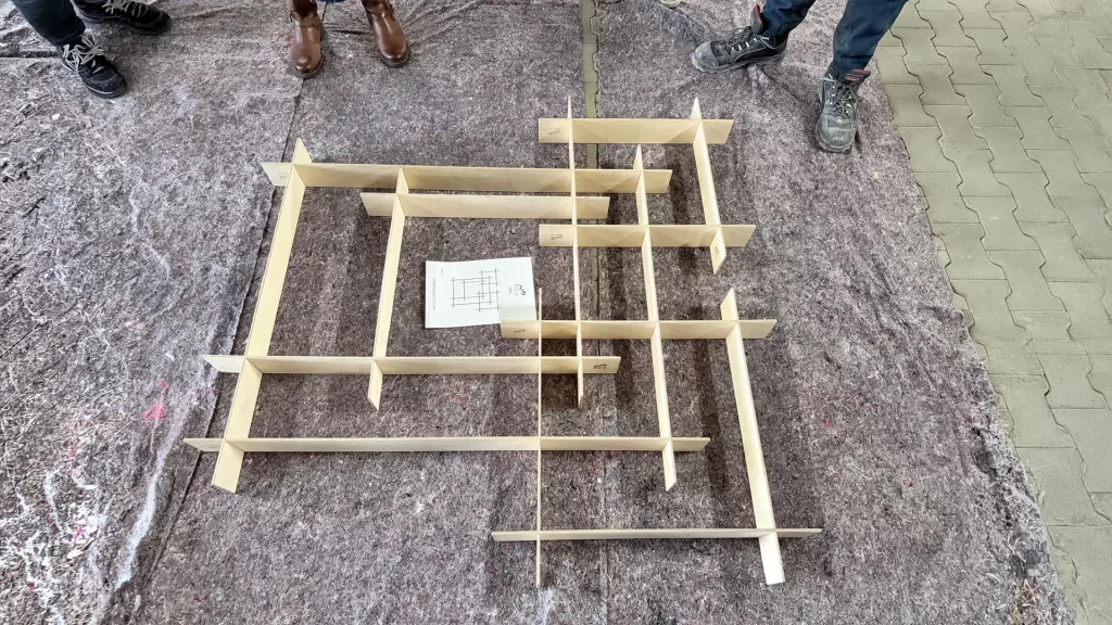 Holz-Elemente werden von mehreren Personen zu einer komplexen Struktur am Boden zusammengesetzt.
