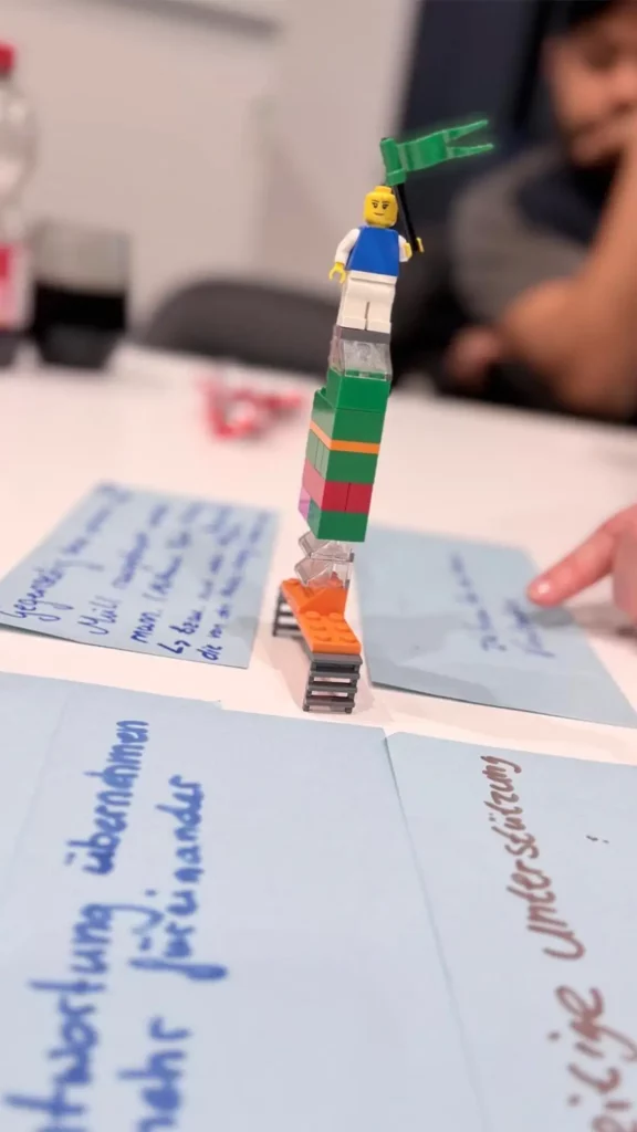 Kleine Lego-Figur auf einem hohen, wackeligen Turm als Symbol für eine Teamaufgabe.