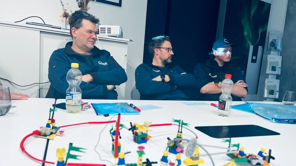 Teilnehmende sitzen mit verschränkten Armen am Tisch und betrachten gebaute Lego-Modelle im Vordergrund.