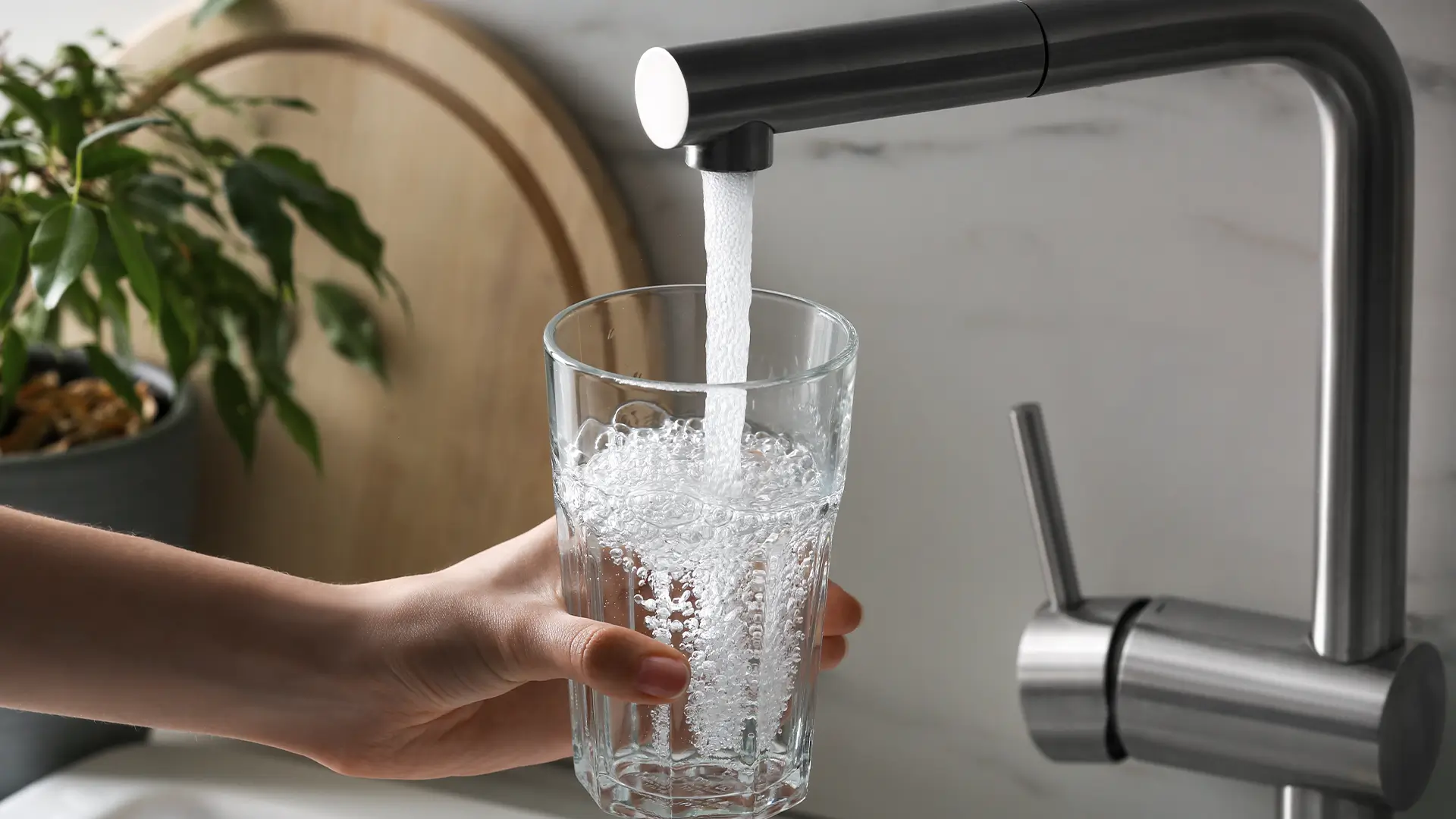 Hand hält ein Glas, das unter einem Wasserhahn mit frischem, sprudelndem Wasser gefüllt wird.