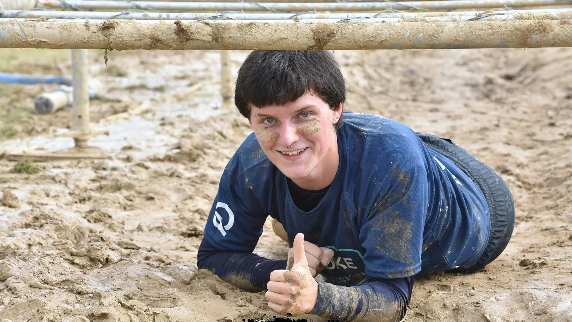Teilnehmerin des Mud Masters liegt im Schlamm unter einem Hindernis und zeigt den Daumen nach oben.